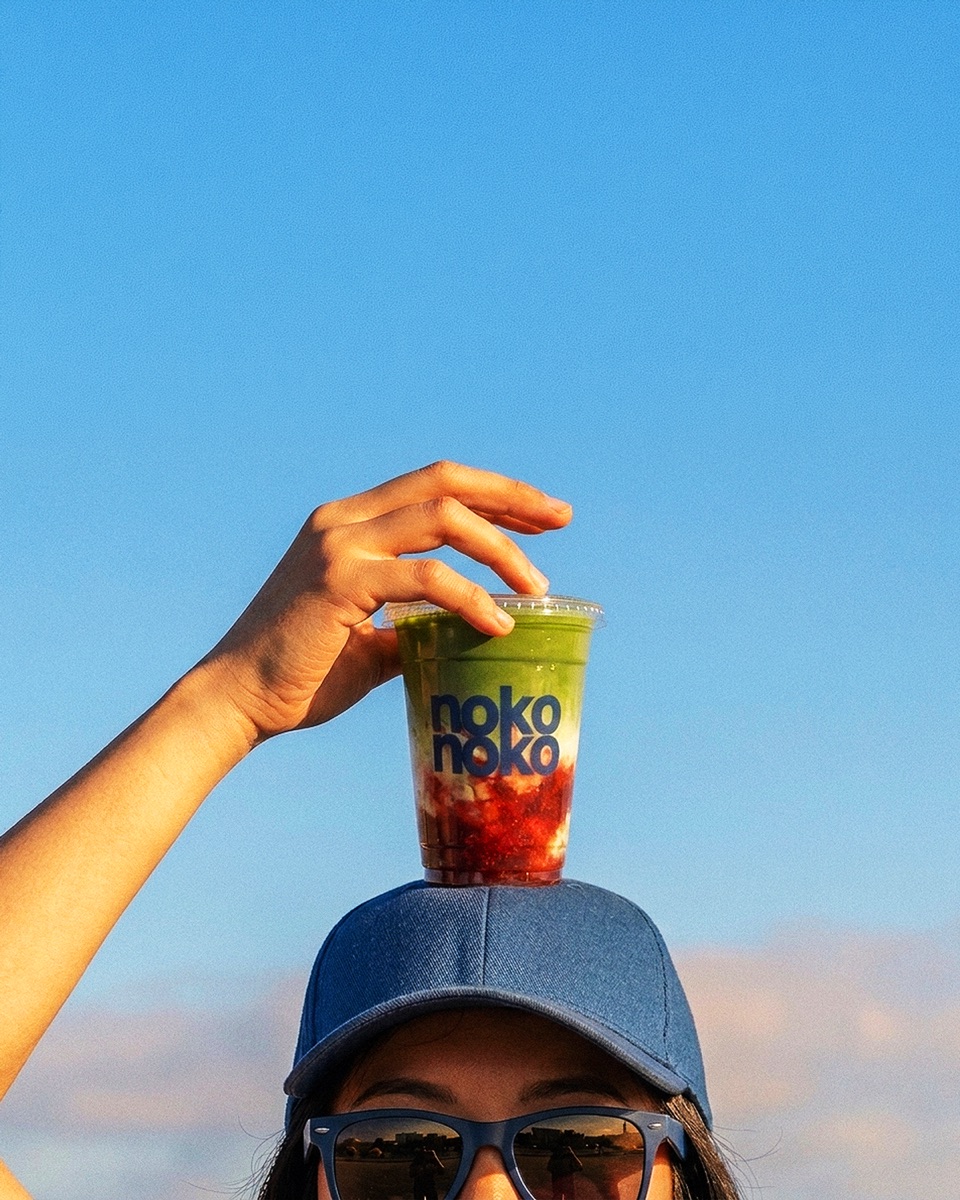 A hand balancing a nokonoko iced strawberry matcha on a denim cap against a blue sky
